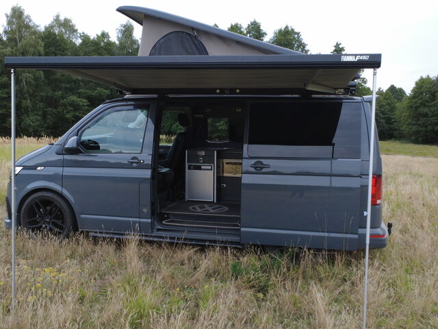 Vincent set up in a meadow with the wind-out side awning extended, sliding door open and galley kitchen visible